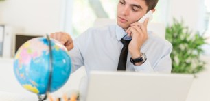 Businessman using phone and looking at globe at discovering available markets of global business. He pointing finger on the globe on the table in front of him.